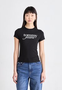 Juodas medvilnės marškinėliai su balta "tommy" logotipo spauda. Trumpomis rankovėmis, prigludusi forma, apvalios iškirptės. Dėvimi su mėlynomis džinsinėmis kelnėmis.