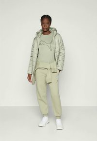 Lys grå dunjakke over en blek grønn genser med logo, sammen med matchende joggebukser og hvite sneakers. Myk, avslappet passform.