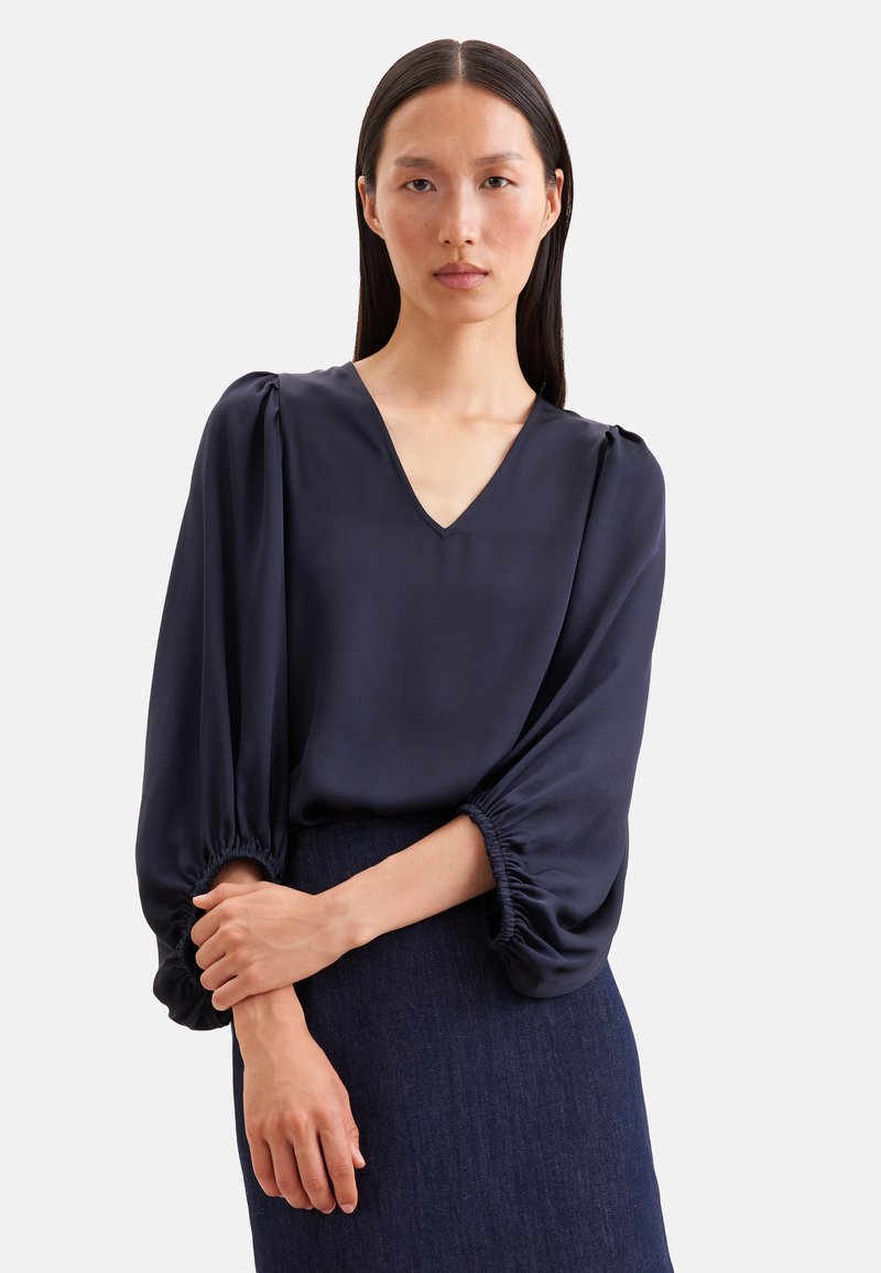 Femme aux longs cheveux foncés porte un chemisier bleu marine foncé à manches bouffantes 3/4 et col en V, associé à une jupe texturée foncée.