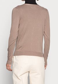 Person som bär en ljusbrun långärmad tröja och vita byxor, sedd från ryggen mot en enfärgad bakgrund.