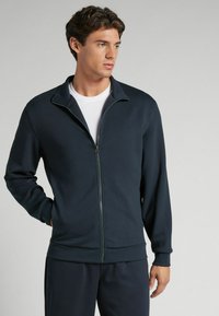 Sudadera navy con cremallera, cuello alto, mangas largas, puños y dobladillo elásticos, hecha de una tela suave, combinada con una camiseta blanca debajo.