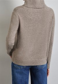 Pull en maille beige avec un col montant, présentant des accents côtelés et une coupe décontractée. Porté avec un jean denim bleu, illustrant un style décontracté.