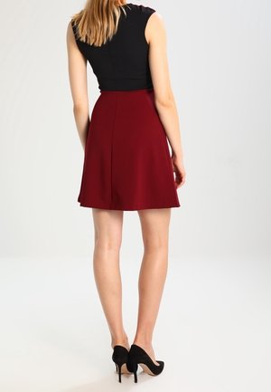 Femme debout de dos, vêtue d'un haut noir sans manches, d'une jupe rouge jusqu'aux genoux et de chaussures à talons noirs, sur un fond uni.