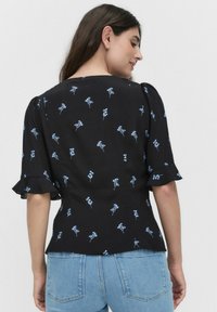 Frau mit langen braunen Haaren, die eine schwarze Bluse mit kleinen blauen Blumenmustern und hellblauen Jeans trägt, mit dem Rücken zu einer schlichten Hintergrundwand.