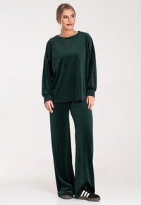 Ciemnozielony komplet dresowy z weluru, składający się z luźnej, długiej koszuli z rękawem oraz spodni z szerokimi nogawkami, charakteryzujący się miękką fakturą i swobodnym krojem. W zestawie czarne trampki.