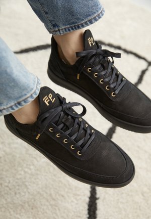 Zwarte leren sneakers met gouden veteroogjes en logo op de tong, gedragen met lichtblauwe spijkerbroek op een licht gemêleerd tapijt.