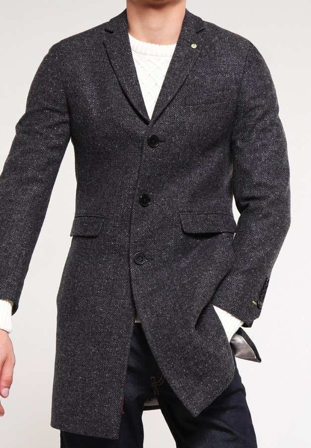 Homme portant un manteau gris foncé à chevrons par-dessus un pull blanc en maille, une main dans la poche du manteau, sur un fond uni.