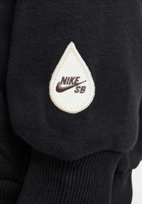 Mâneca unui hanorac negru cu un patch din material alb în formă de lacrimă și logo-ul "NIKE SB" cusut pe acesta, de culoare maro închis.