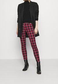 Femme portant un pantalon ajusté en tartan rouge et noir, un col roulé noir, un long blazer noir et des bottines noires sur un fond uni.