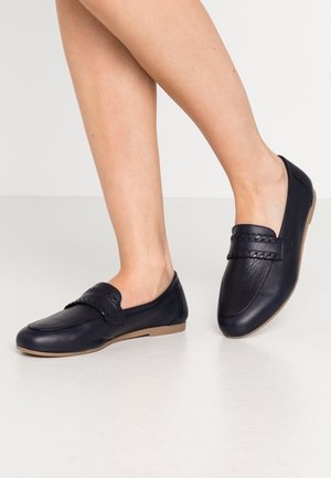 Jambes portant des mocassins en cuir noir à enfiler avec un détail de sangle tressée et des semelles plates marron clair sur une surface blanche lisse.
