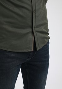 Dunkelgrünes, langärmliges Hemd mit Knopfleiste, das eine glatte Textur und einen gespaltenen Saum aufweist, kombiniert mit dunkelblauen Jeans.