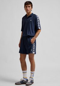 Navy polo och shorts-set i strukturerad tyg, med vita chevron-accenter på ärmarna och sidan. Grå sneakers och randiga strumpor kompletterar looken.