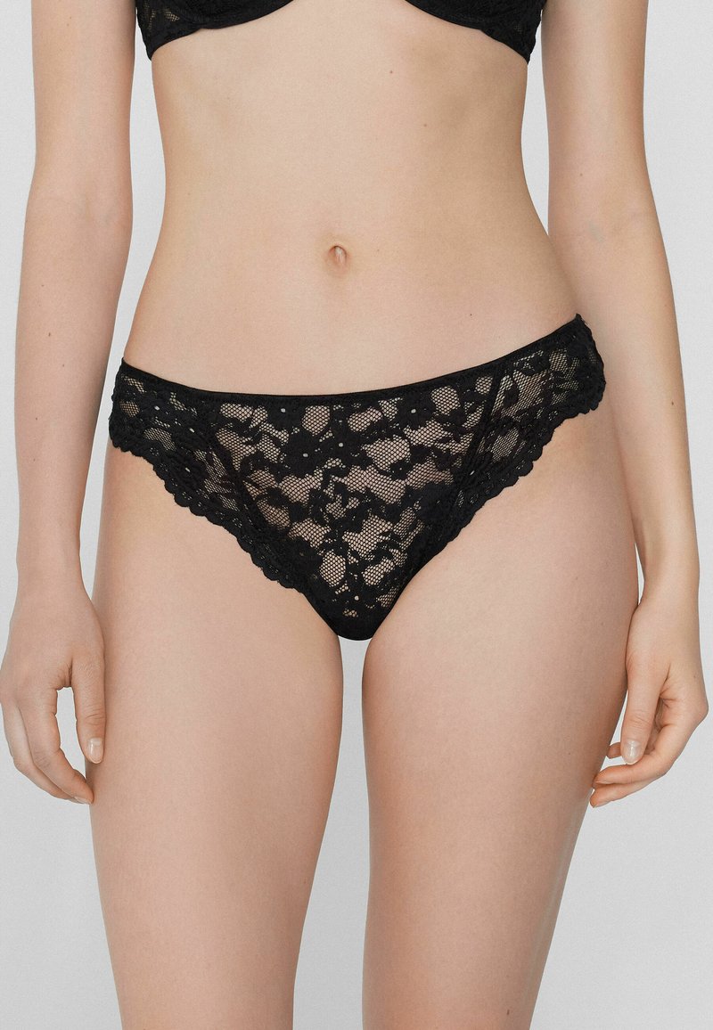 String noir en dentelle avec motif floral, bords festonnés et texture transparente, doté d'une taille mi-haute et d'un design minimaliste.