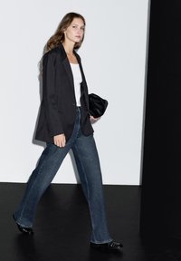 Černé sako přes bílý top, v kombinaci s tmavě modrými širokými džíny a černou obuví. Modelka drží černé psaníčko a kráčí proti jednoduchému pozadí.