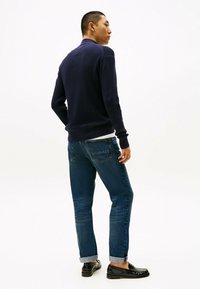 Homme en pull bleu marine, jeans bleus retroussés et mocassins noirs, se tenant avec le dos partiellement tourné contre un fond blanc uni.