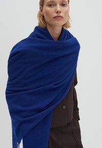 Sciarpa di lana blu dalla texture morbida, con un drappeggio fluido e bordi sfrangiati. Indossata sopra un blazer marrone scuro.