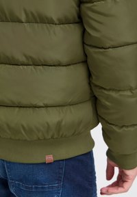 Olivgrüne Steppjacke mit horizontaler Steppung, gerippten Bündchen und einem kleinen braunen Etikett am Saum. Sie hat eine glatte, glänzende Textur.