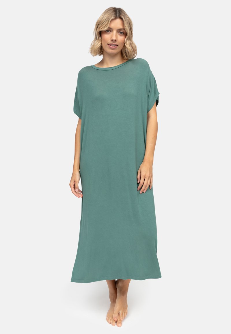 Frau steht barfuß und trägt ein lockeres, knöchellanges, kurzärmeliges grünes Kleid vor einem schlichten weißen Hintergrund.