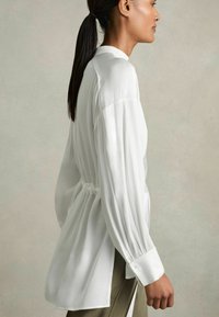 Camicia bianca a maniche lunghe con texture arricciata e vita con cintura, dotata di polsini con bottoni e una silhouette rilassata. Tessuto leggero e liscio.