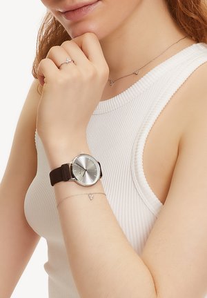 Runde Silberuhr mit einem braunen Silikonarmband, ausgestattet mit einem schlanken grauen Zifferblatt. Begleitet von einem zarten Silberarmband mit kleinen Akzenten.