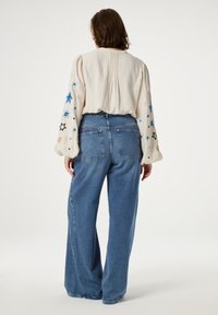 Lichtbeige blouse met pouf mouwen en geborduurde sterren in blauw en zwart, gecombineerd met losse blauwe denim jeans.