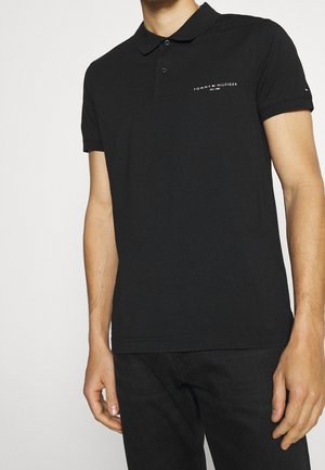 Mężczyzna w czarnej koszulce polo Tommy Hilfiger z logo na piersi i rękawie, w połączeniu z czarnymi spodniami, stoi na tle jednolitym.