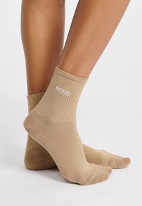 Calcetines de color beige con canalé y un sutil brillo, que presentan un logo blanco de "BOSS" en el puño. De altura tobilleras con textura suave.
