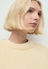 Suéter de punto suave en color amarillo con un cuello redondo acanalado, llevado sobre una camisa blanca, que presenta una tela texturizada y acogedora.