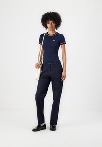 Marineblå t-skjorte med korte ermer og røde og hvite kanter, paret med marineblå bukser og svarte loafers; modellen holder en kremfarget totebag.