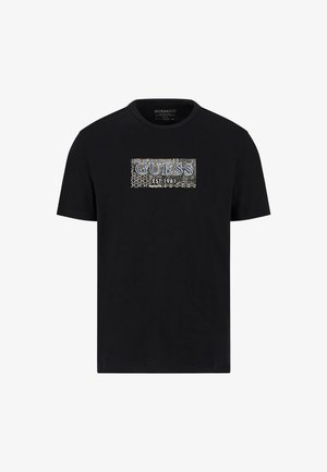 Black cotton t-shirt featuring a rectangular graphic with "GUESS" and "EST. 1981" in blue, surrounded by a patterned border. Round neck design.