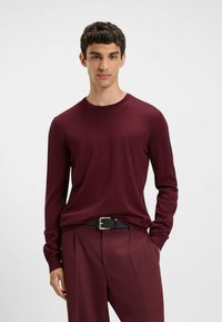 Burgunderfarbener Pullover mit langen Ärmeln und Rundhalsausschnitt, aus weichem Material. Kombiniert mit maßgeschneiderten burgunderfarbenen Hosen und einem schwarzen Gürtel mit einer silbernen Schnalle.