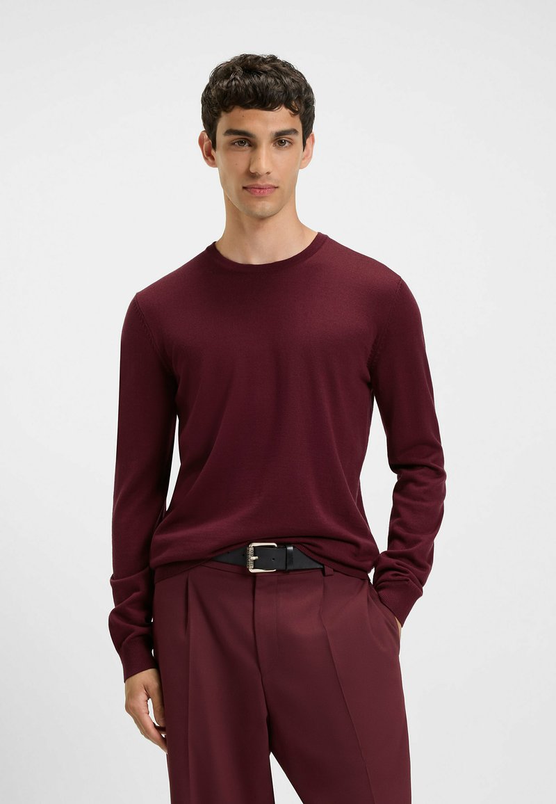 Burgunderfarbener Pullover mit langen Ärmeln und Rundhalsausschnitt, aus weichem Material. Kombiniert mit maßgeschneiderten burgunderfarbenen Hosen und einem schwarzen Gürtel mit einer silbernen Schnalle.