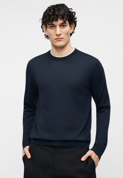 Homme aux cheveux bouclés et foncés portant un pull bleu marine à col rond et un pantalon noir, les mains dans les poches, faisant face à l'avant.