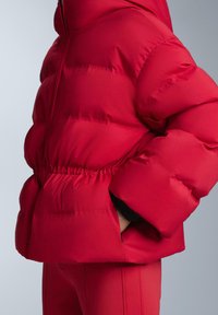 Piros steppelt dzseki, melynek derékrésze összehúzott, ujjai pedig túlméretezettek. Simább, fényes anyagból készült, steppelt tapintással és oldalzsebekkel.