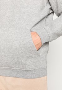 Felpa grigia con una texture morbida, tasca a marsupio e polsini a coste. Abbinata a pantaloni di colore chiaro.