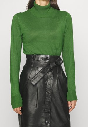 Mujer con un suéter de cuello alto verde metido en una falda de cuero negra de cintura alta, con un cinturón anudado y botones en la parte delantera.