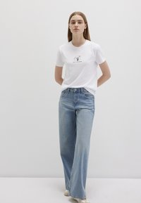 Weißes Baumwoll-T-Shirt mit einem grafischen Druck auf der Brust. Kombiniert mit hellblauen, weiten Jeans. Das Modell steht vor einem schlichten Hintergrund.