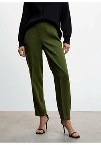 Pantalon taillé vert avec une texture lisse, présentant des plis sur le devant et une taille ajustée, associé à des escarpins noirs à lanières.