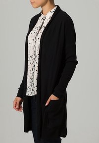 Persona che indossa un cardigan aperto nero su una camicia bianca con macchie nere e grigie, mano nella tasca del cardigan.