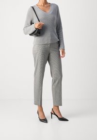Une femme portant un pull en tricot gris, un pantalon à carreaux gris jusqu'aux chevilles, des escarpins à bride arrière noirs, et tenant un petit sac à bandoulière noir.