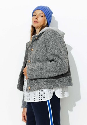 Veste grise en mélange de laine avec un design court, des boutons dorés et une capuche grise. Un top en broderie blanche et un pantalon marine avec des rayures bleues sont portés en dessous.