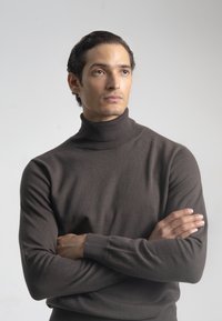 Hombre con los brazos cruzados, vistiendo un suéter de cuello alto gris oscuro, mirando levemente de lado contra un fondo claro y liso.