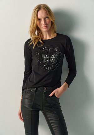 Mujer con pelo largo y rubio lleva una camiseta negra de manga larga con diseño de mariposa y pantalones negros ajustados, y está de pie contra una pared gris claro.