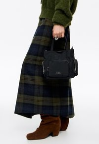 Osoba trzymająca małą czarną torebkę z kieszeniami na zamek z przodu i z boku, ubrana w zielony miękki sweter, niebiesko-zieloną spódnicę w kratę oraz brązowe zamszowe kozaki.