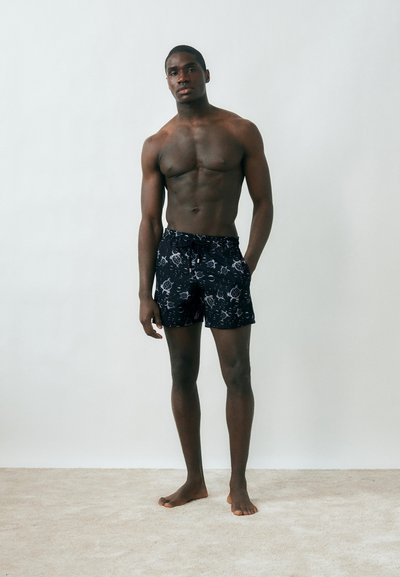 Άνδρας χωρίς πουκάμισο, όρθιος, ξυπόλητος, ντυμένος με μαύρο μαγιό με λευκά σχέδια θαλάσσιας χελώνας και κυμάτων, πάνω σε απλό φωτεινό φόντο.