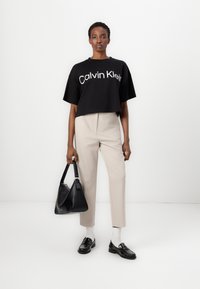 Black cropped t-shirt with white lettering, beige high-waisted trousers, black bag, and black loafers with white socks. Minimalist design.