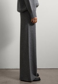 Personne portant une longue jupe en tricot gris, un pull en tricot gris assorti et des mocassins noirs, se tenant devant un mur léger uni.