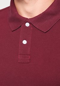 Polo shirt bourgogne avec un col côtelé, deux boutons blancs et un tissu texturé. Design simple et classique avec un ourlet droit.