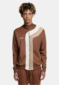 Brun zip-up jakke med høy krage, med hvite og beige geometriske paneler og en liten logo på venstre bryst. Glatt stofftekstur.