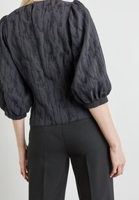 Blusa grigia scura con maniche a sbuffo, realizzata in un tessuto testurizzato con un sottile motivo ondulato. Chiusura sul retro del collo visibile, indossata con pantaloni neri.